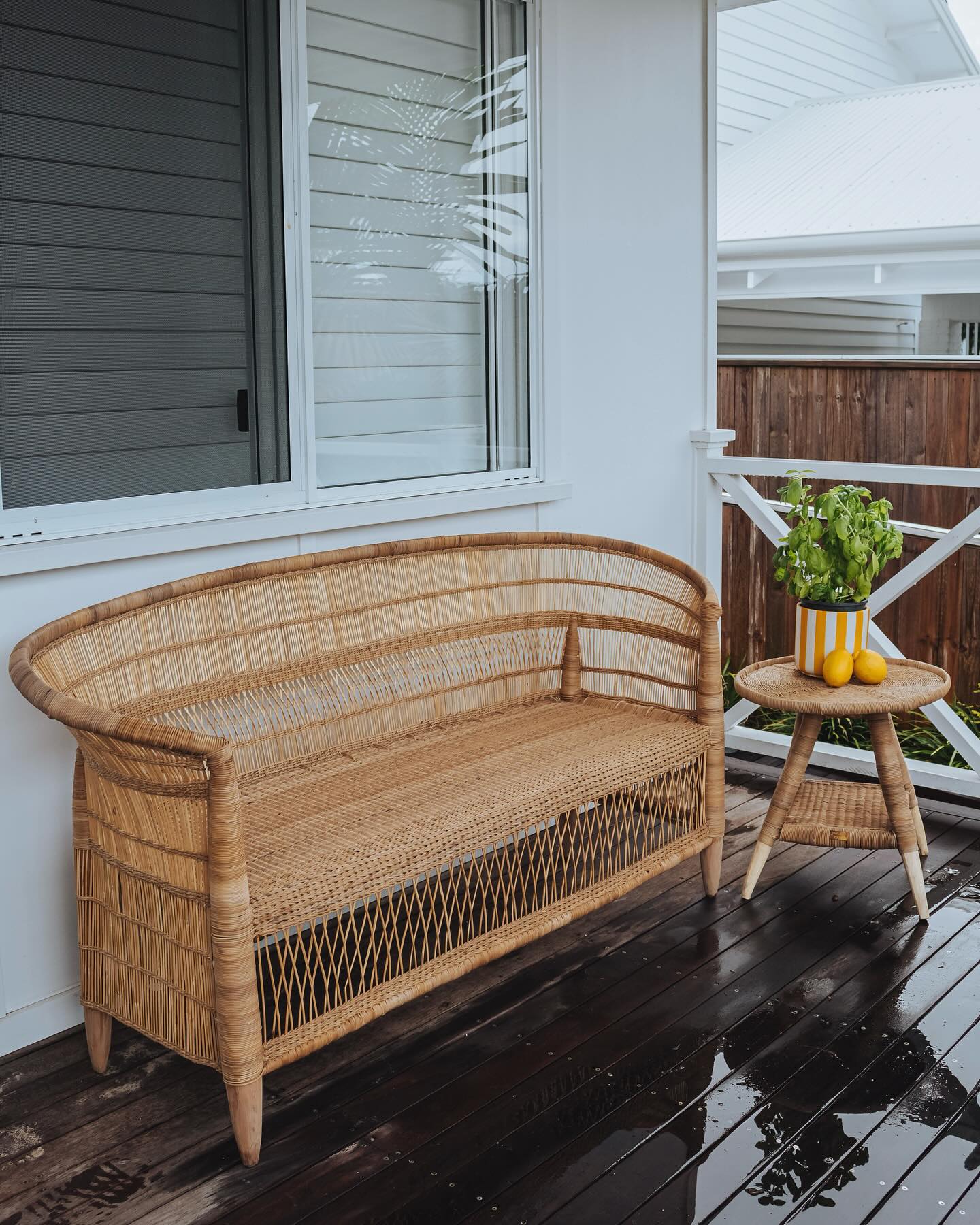 Wicker bench on a wooden deck with a small table and plants in the background.