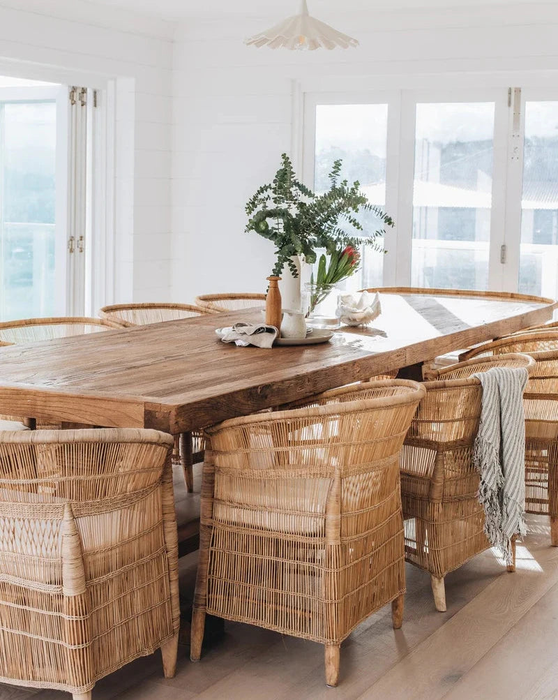 A set of natural color Malawi cane chairs with a round wooden table, placed in a bright room with a vase of flowers on the table and a light hanging above.