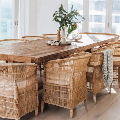 A set of natural color Malawi cane chairs with a round wooden table, placed in a bright room with a vase of flowers on the table and a light hanging above.