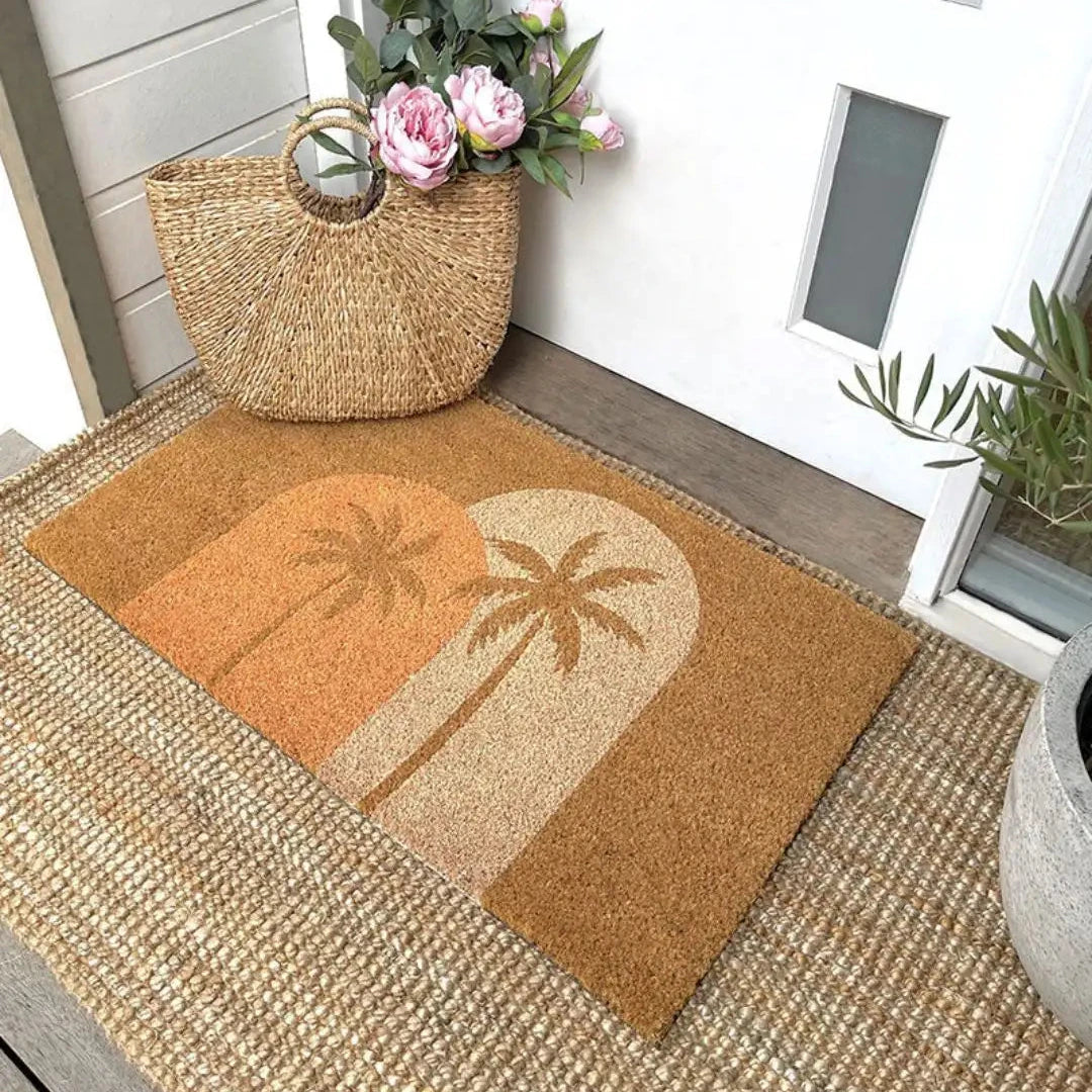 Doormat with palm tree design on a wooden floor, next to a woven bag and flowers.