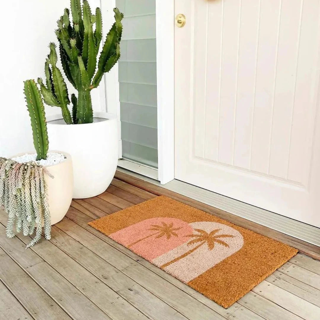 Doormat with palm tree design on a wooden deck with potted plants.