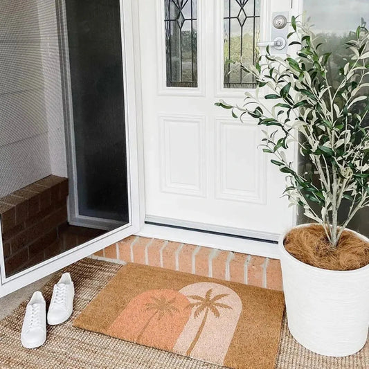Front door with a doormat featuring palm trees, a plant in a pot, and white shoes on a rug.