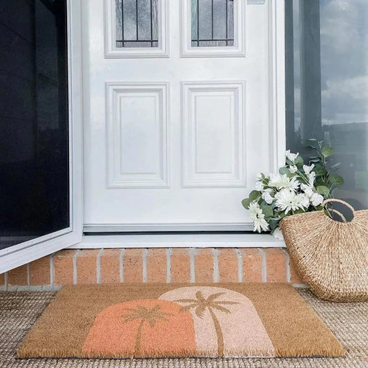 White front door with decorative glass panels, doormat with palm tree design, and a woven basket on a brick porch.