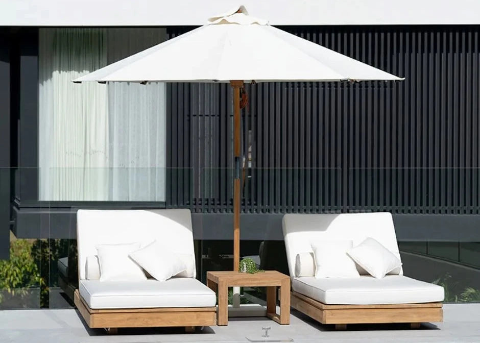 Two white cushioned lounge chairs with a wooden table and umbrella by a poolside.