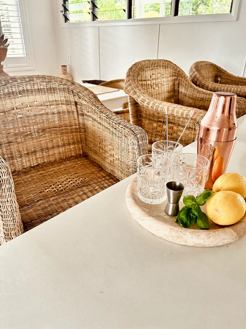 Cocktail shaker, glasses, and fruits on a table with wicker stools in the background