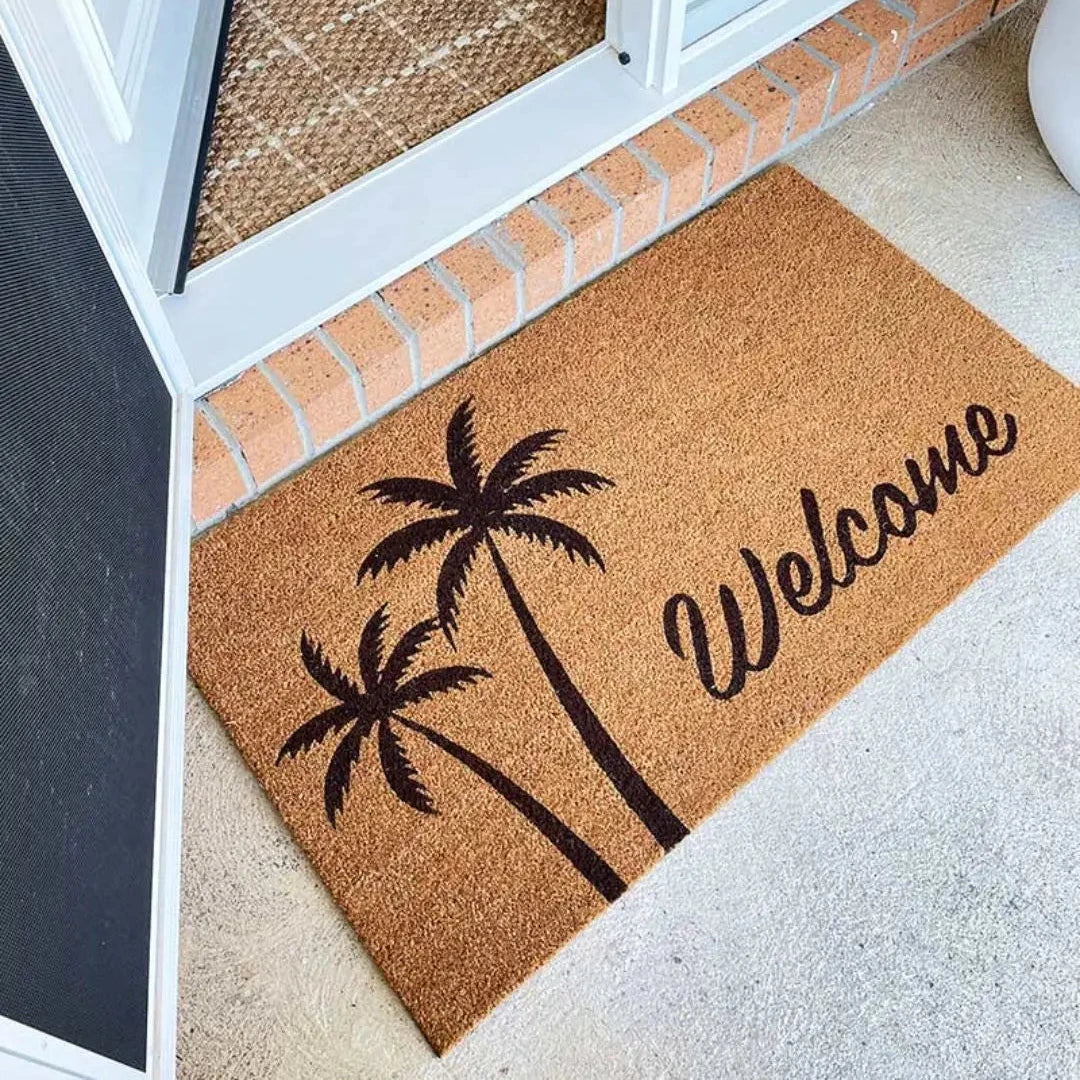 Doormat with palm tree design and 'Welcome' text on a carpeted floor.