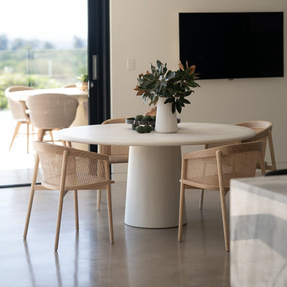 Modern dining area with a round white table and wicker chairs in a bright room. Featuring the Elio Round Microcement Dining Table – Seashell Finish from Sun Republic