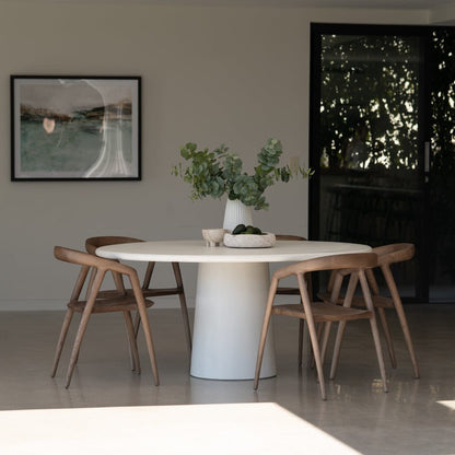 Dining area with a white round table and wooden chairs, featuring a vase with greenery on the table. Featuring the Elio Round Microcement Dining Table – Seashell Finish from Sun Republic