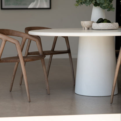Dining area with a white round table and wooden chairs, featuring a vase with greenery on the table. Featuring the Elio Round Microcement Dining Table – Seashell Finish from Sun Republic