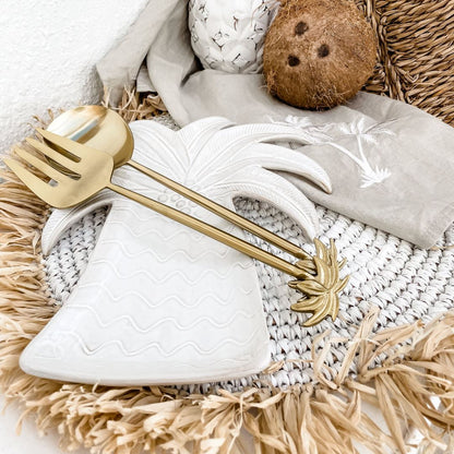 A pair of gold brass palm tree salad servers, consisting of a spoon and fork, displayed on a white napkin with a decorative pineapple and coconut in the background.