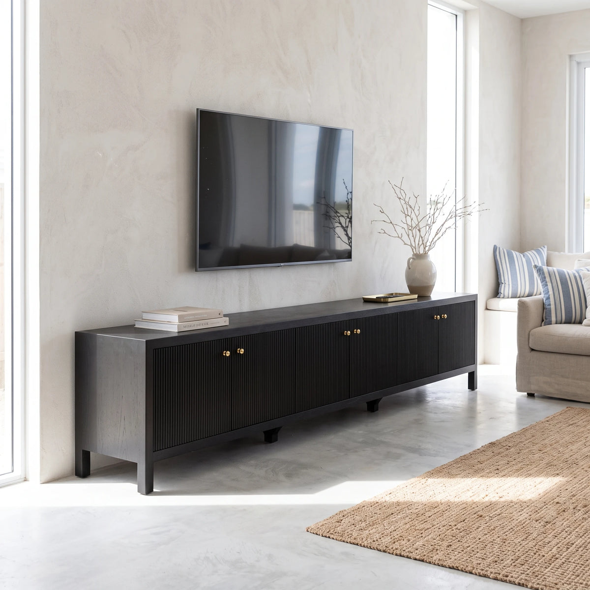 A long black TV console with ribbed front doors and small gold knobs, placed against a light textured wall in a bright living room. A wall-mounted TV is above the console, with books and a vase on top, and part of a sofa and rug are visible nearby.