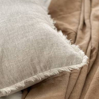 A close-up of a light beige linen pillow with frayed edges resting on a brown blanket over white bedding.
