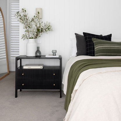 A modern black ribbed nightstand in a styled bedroom setting, featuring a white vase with greenery, a gray bottle, and books on its top shelf, with a bed dressed in white and olive green textiles beside it.