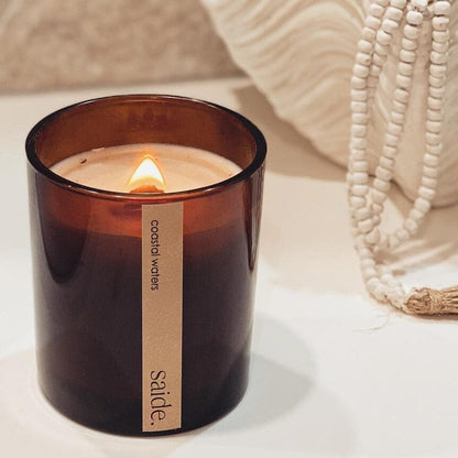 Candle in a brown glass jar with a label, placed on a light surface with a blurred background.
