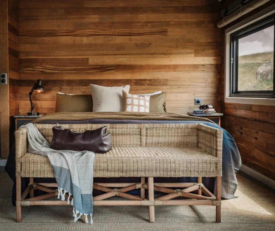 Wooden bench with a blanket and bag in front of a wooden wall with a bed and lamp.