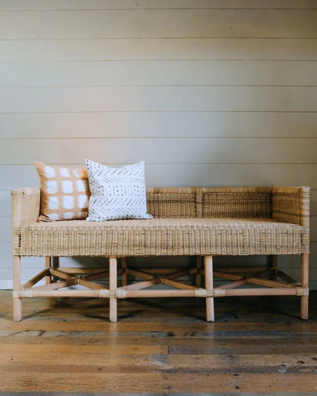 Wicker bench with two pillows against a wooden wall.