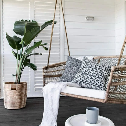 Wicker swing with cushions on a wooden deck, next to a potted plant and small table with a cup.