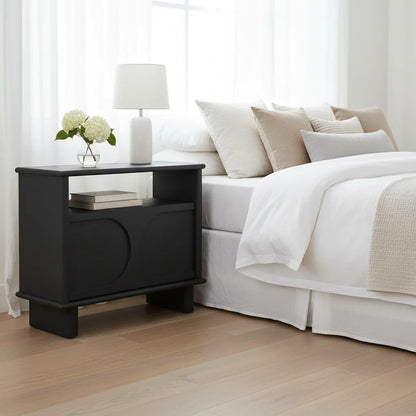 Black nightstand with white lamp and books next to a bed with beige and white bedding.