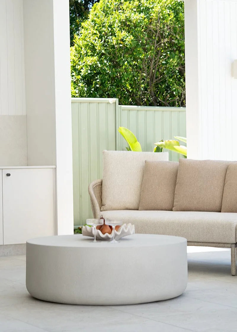 Beige outdoor sofa with a round coffee table on a patio with greenery in the background