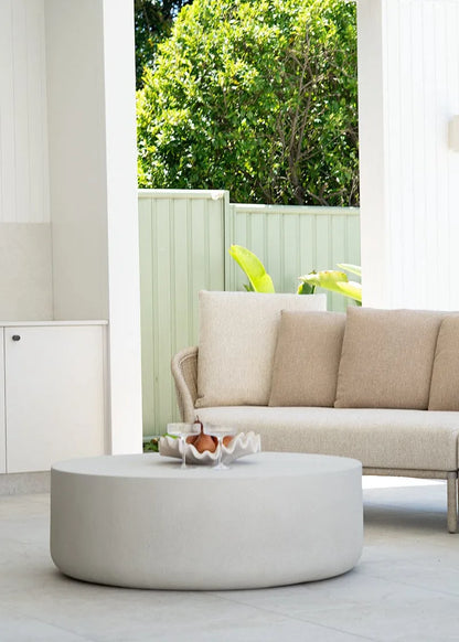 Beige outdoor sofa with a round coffee table on a patio with greenery in the background