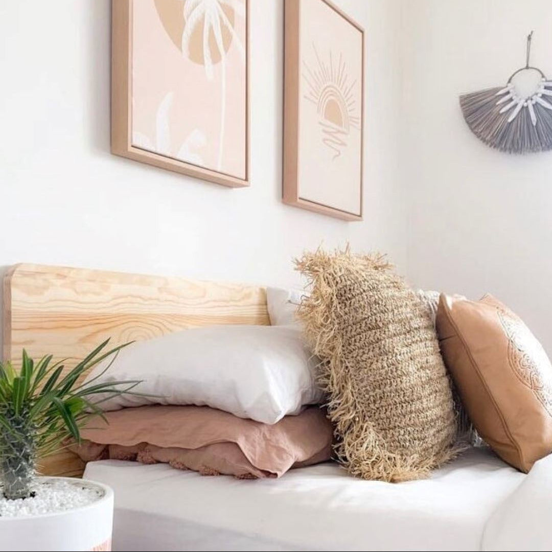 A styled bedroom corner with two framed minimalist wall art prints above a bed. The prints feature pale peach and beige tropical sun and palm designs in light wood frames.