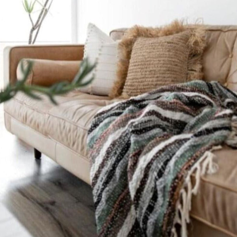A cozy beige leather sofa in a bright living room setting, styled with two textured throw pillows and a fringed multicolor knit blanket draped over the seat.