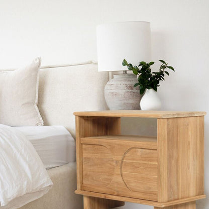 Wooden nightstand with lamp and plant next to a bed in a bedroom setting