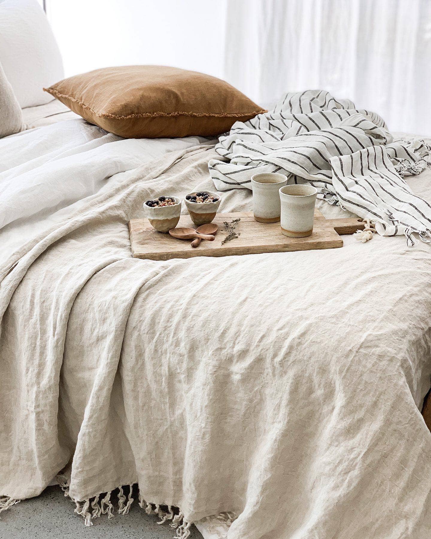 Bed with beige bedding, brown pillow, and small wooden tray with items on a light background