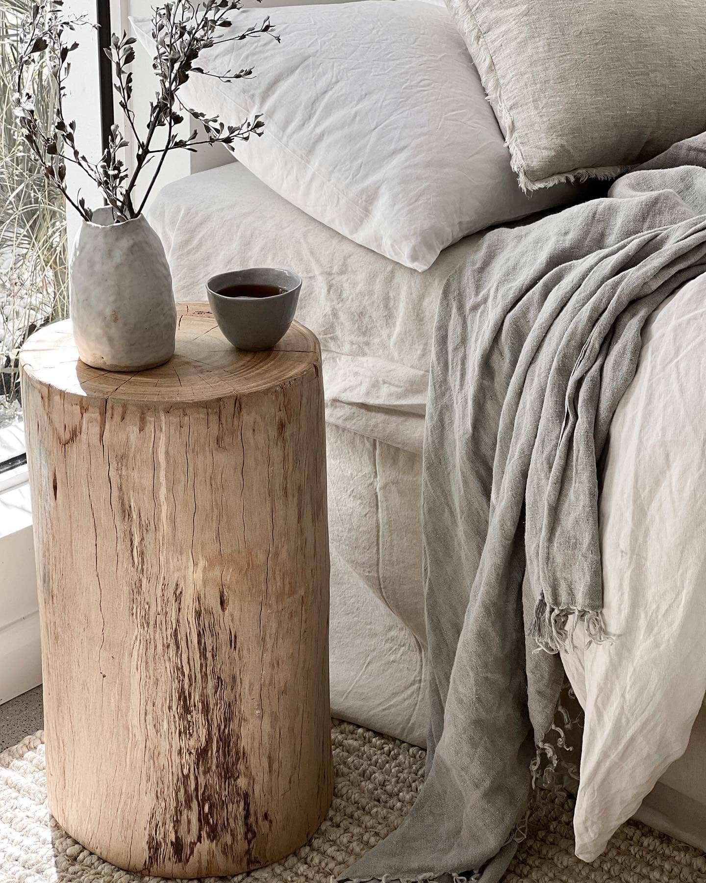 Wooden side table with a vase and cup next to a bed with white bedding and gray blanket.