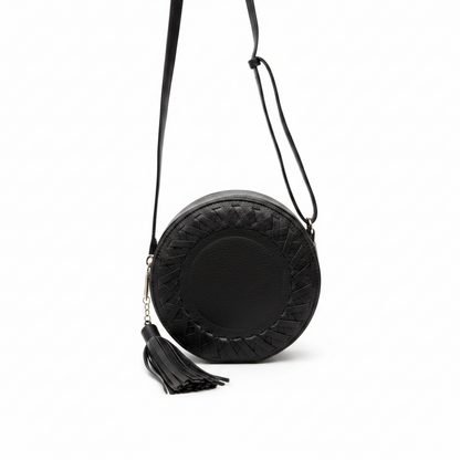 A black round crossbody bag with a textured quilted border, a central pebbled leather panel, a hanging black tassel charm with a small gold chain, and a thin black shoulder strap, shown on a plain white background.