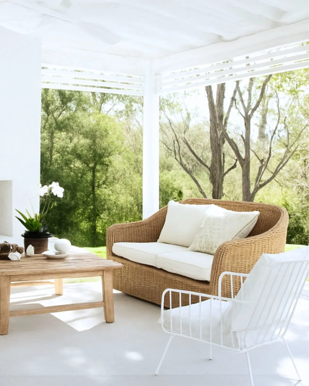 Outdoor patio with wicker sofa, coffee table, and white chairs with a view of trees.