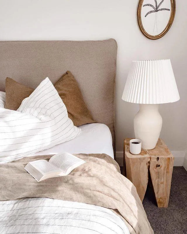 Neatly made bed with pillows and a lamp on a teak peg stool side table in a bedroom.