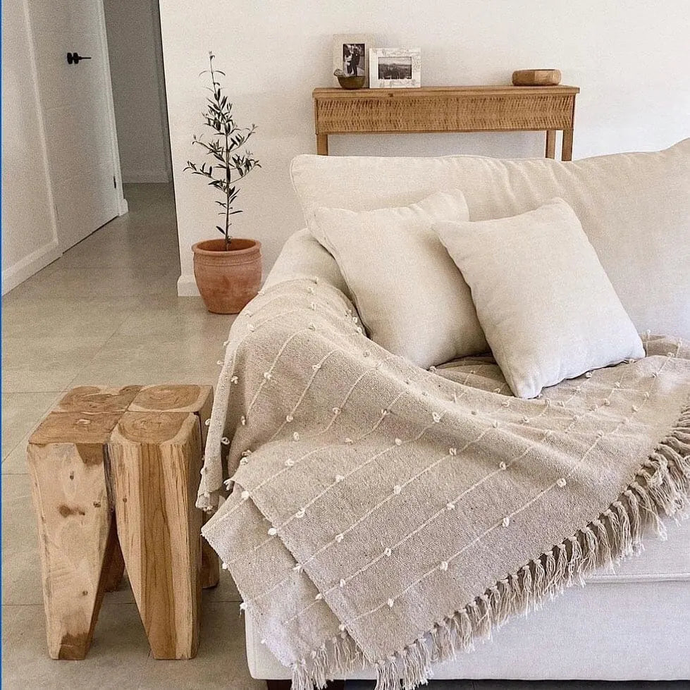Beige textured blanket draped over a white sofa with teak peg stool and a plant in the background.