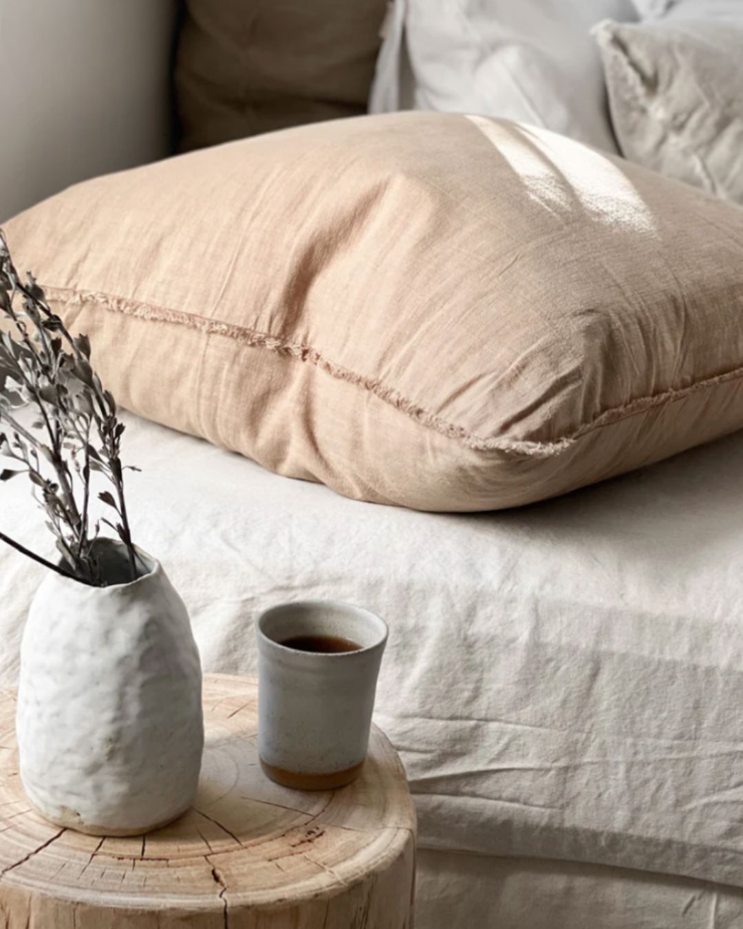 Beige pillow on a bed with a small wooden table holding a cup and vase.