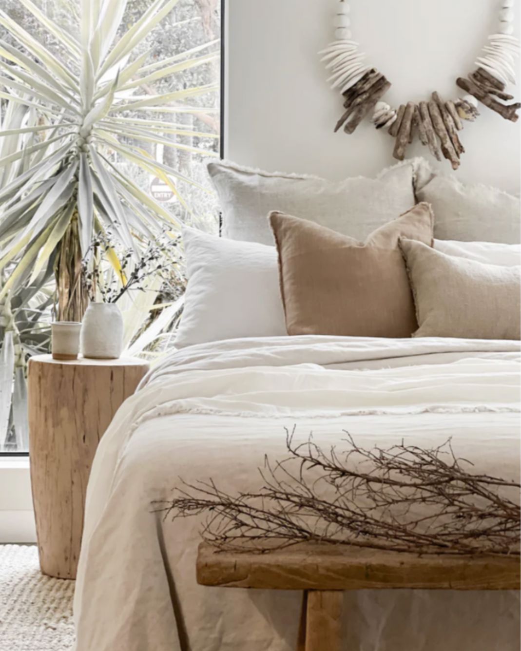 Neatly made bed with beige and white pillows, wooden bench, and decorative wreath in a bright room.