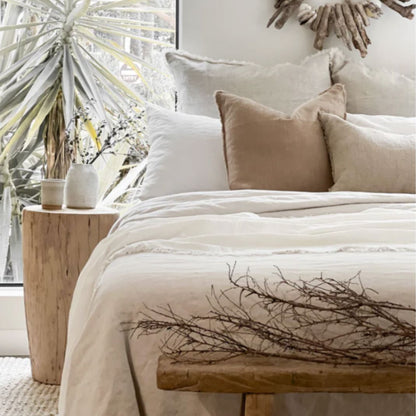 Neatly made bed with beige and white pillows, wooden bench, and decorative wreath in a bright room.