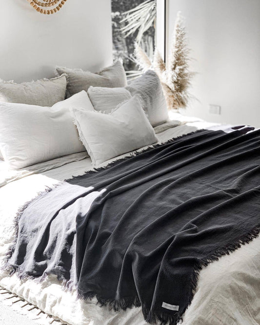 Neatly made bed with white pillows and a dark gray throw blanket in a bright room.