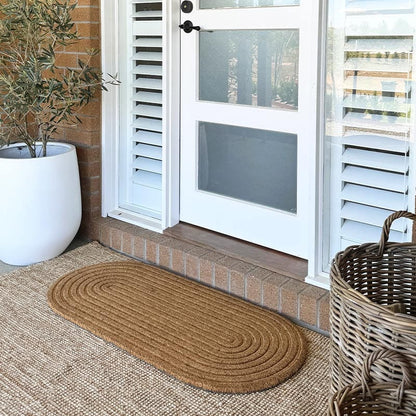 A large oval-shaped coir doormat with a ribbed texture placed on a doorstep, leading up to a white door with glass panels from Sun Republic