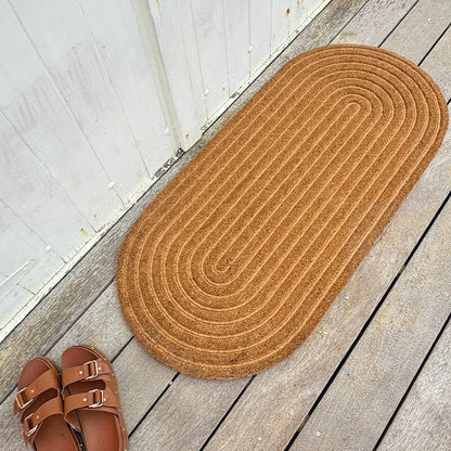 A large oval-shaped coir doormat with a ribbed texture placed on a doorstep, from Sun Republic