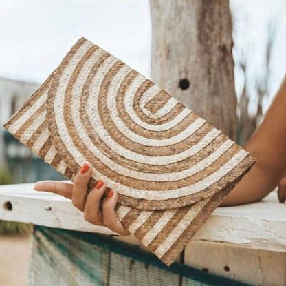 A person holds a woven rectangular clutch or envelope-style bag with a spiral stripe pattern in natural tan and off-white. The photo is taken outdoors near a wooden railing and tree trunk, with the person's hand and arm visible.
