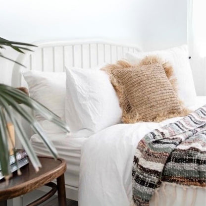 A styled bedroom scene showing a bed with white bedding, a textured beige throw pillow, and a multicolor striped woven blanket draped over the corner. A dark wood side table with a plant is visible in the foreground.