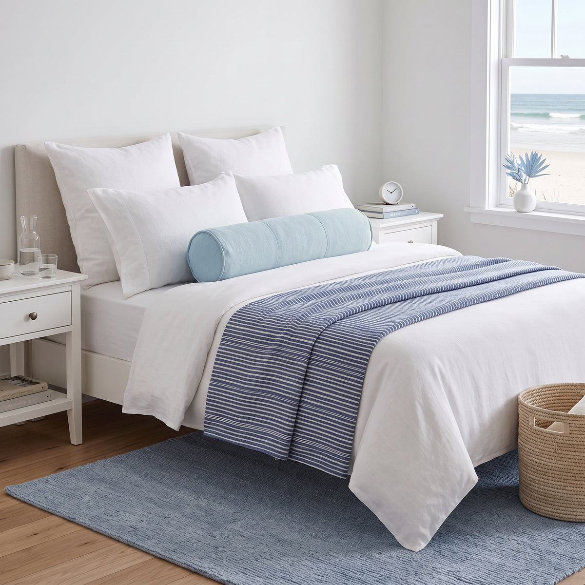 A neatly made bed in a bright bedroom setting, styled with white bedding, a light blue cylindrical bolster pillow, and a blue-and-white striped throw blanket. The room includes white nightstands, a window with a beach view, a blue rug, and a woven basket.
