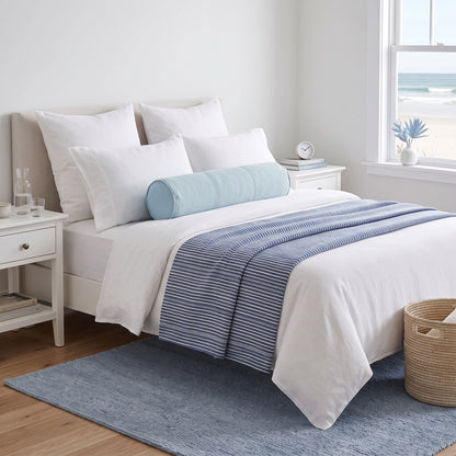 A neatly made bed in a bright bedroom setting, styled with white bedding, a light blue cylindrical bolster pillow, and a blue-and-white striped throw blanket. The room includes white nightstands, a window with a beach view, a blue rug, and a woven basket.