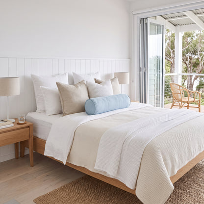 A neatly made bed with white and cream bedding, beige pillows, and a light blue cylindrical bolster pillow, set in a bright, modern bedroom with a wooden headboard and nightstand.