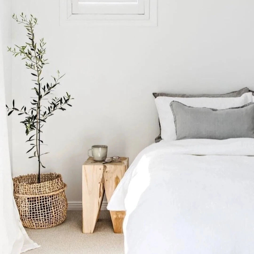 Neatly made bed with gray pillows, wooden side table with mug, and plant in basket in a bright bedroom.