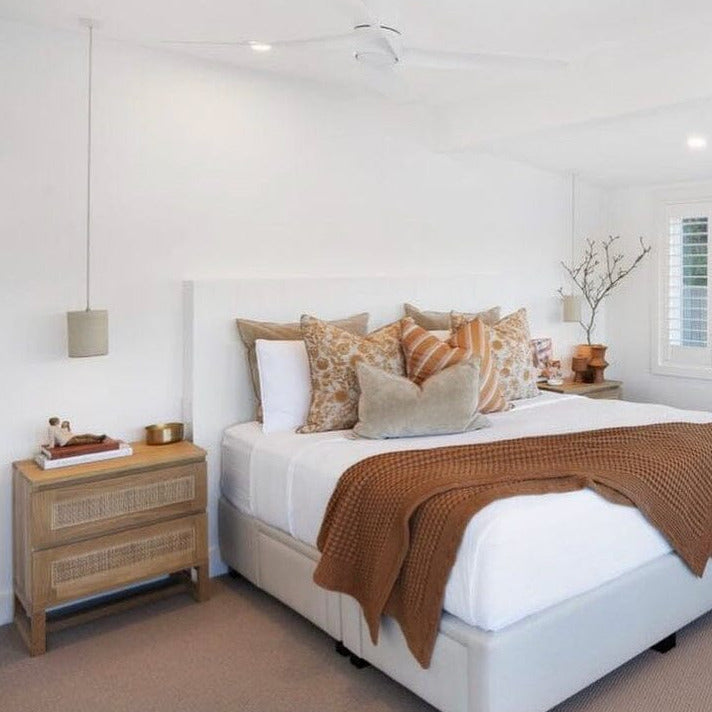 The image shows a Kiera American Oak Nightstand with two drawers, a lamp on top, and a woven rattan panel on the drawer front, placed in a bedroom setting.