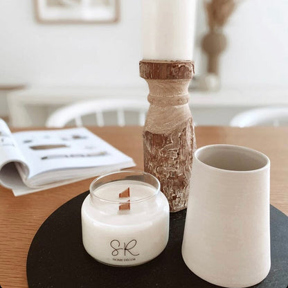 Candle with 'SR' branding on a table with a vase and newspaper in the background