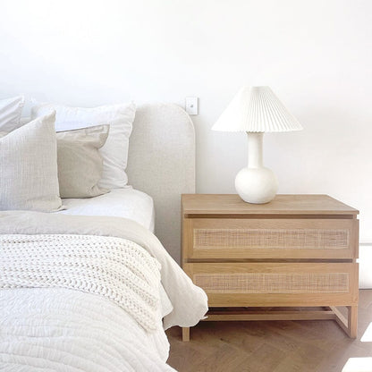 The image shows a Kiera American Oak Nightstand with two drawers, a lamp on top, and a woven rattan panel on the drawer front, placed in a bedroom setting.