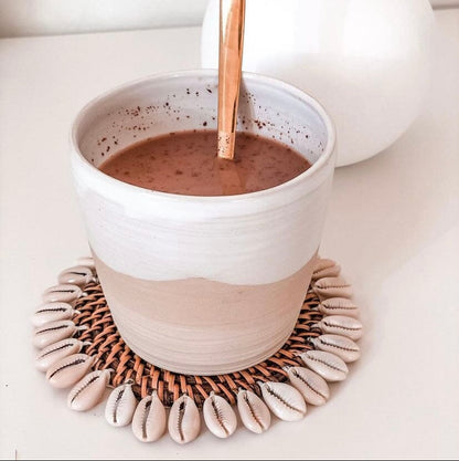 A beige natural rattan coaster with a cowrie shell border, resting on a white surface, with a cup of coffee and a decorative pumpkin in the background.