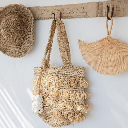 A woven straw tote bag with long fringe and a white tassel hangs from a carved wooden wall hook. A woven straw hat is on the left hook and a woven straw sun hat is on the right hook, all against a white wall.